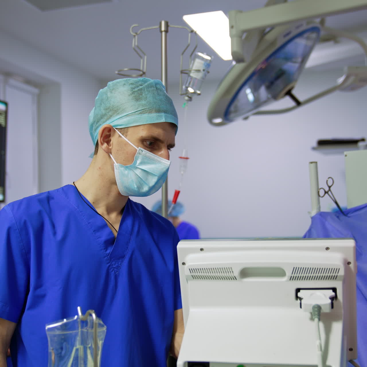 Male medical assistant wearing blue uniform, mask and cap looking intently at monitor. Hospital room for neurosurgical operations setting.