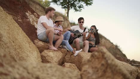 group of young cheerful people sitting on the rocks by the seashore and playing guitar, singing songs and dancing. slow motion