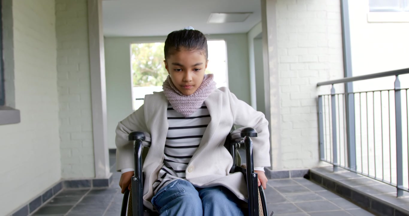 Teen African-American girl grasping rims and pushing wheelchair along covered walkway toward window