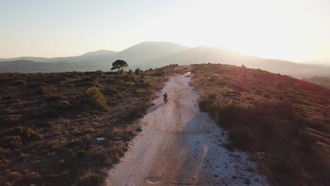 Toma-Aérea-De-Seguir-A-Un-Motociclista-Mientras-Conduce-Por-Un-Camino-De-Ripio-Durante-La-Hora-Dorada-En-Una-Montaña-Al-Lado-De-Atenas,-Grecia