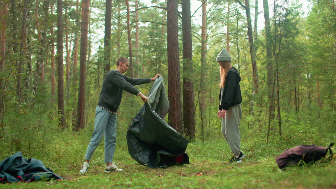 Man lifts and adjusts camping gear while colleague watches attentively in calm forest setting, surrounded by trees and scattered bags on grassy ground, preparing to set up for outdoor camping