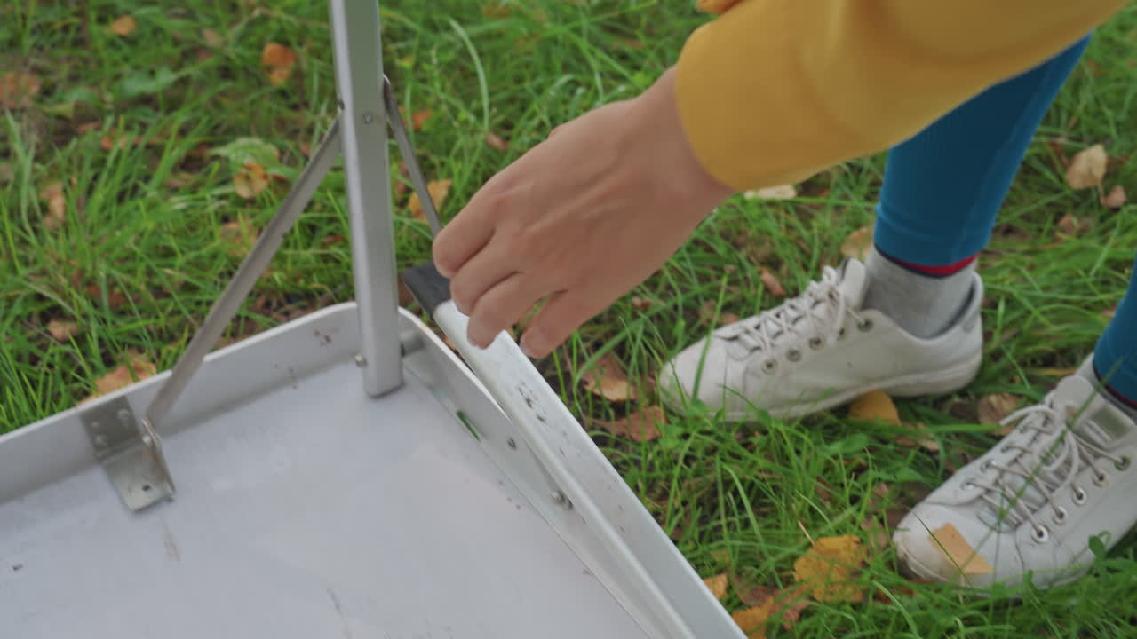 Inspecting camping table final setup on grass, camper checks leg stability and balance, kneeling and leaning over table, casual jacket and leggings visible, satisfied check before picnic