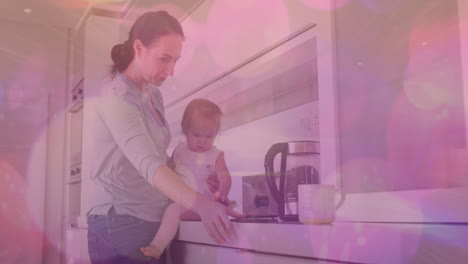 animation of pink spots of light over caucasian woman with baby in kitchen