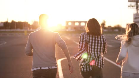 Cerca-De-Cuatro-Amigos-Con-Estilo-Corren-Por-Una-Zona-De-Estacionamiento-Vacía-Al-Aire-Libre-Felicidad-Divertida,-Hombres-Y-Mujeres-Jóvenes-Corren-Al-Atardecer-Y-Luego-Saltan-Juntos-Libremente