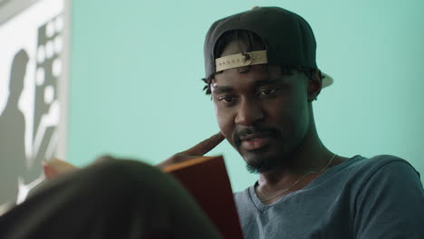 close up of focused freelancer wearing cap reading book with thoughtful expression as bright sunlight enters room creating lens flare and soft glow effect against teal background
