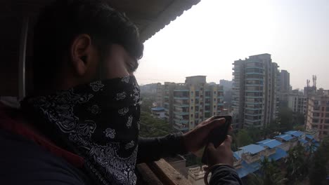 Junge-Mit-Handy-Auf-Terrasse-Waring-Mask-Indien-Mit-Blick-Auf-Gebäude-Neu-Normal