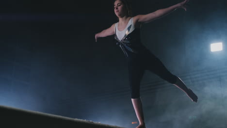 The-girl-is-a-professional-athlete-performs-gymnastic-acrobatic-trick-on-a-beam-in-backlight-and-slow-motion-in-sports-gymnastic-clothing.-Smoke-and-blue.-Jump-and-spin-on-the-balance-beam