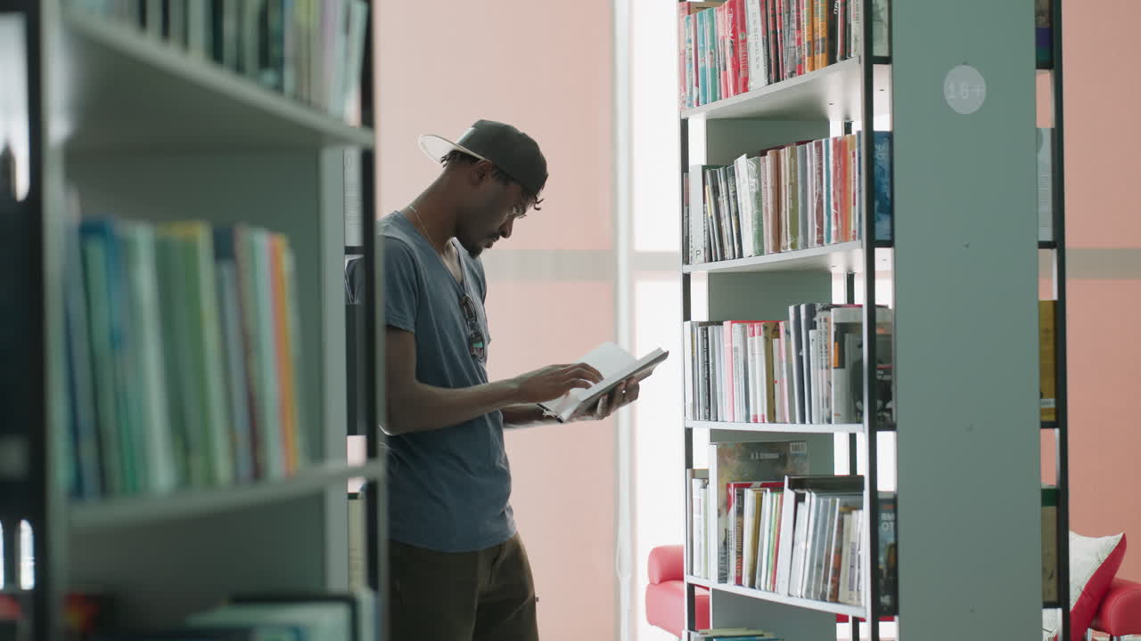 man in casual cap stands between library shelves, reading book with focused expression, turning pages near window light; quiet study room mood