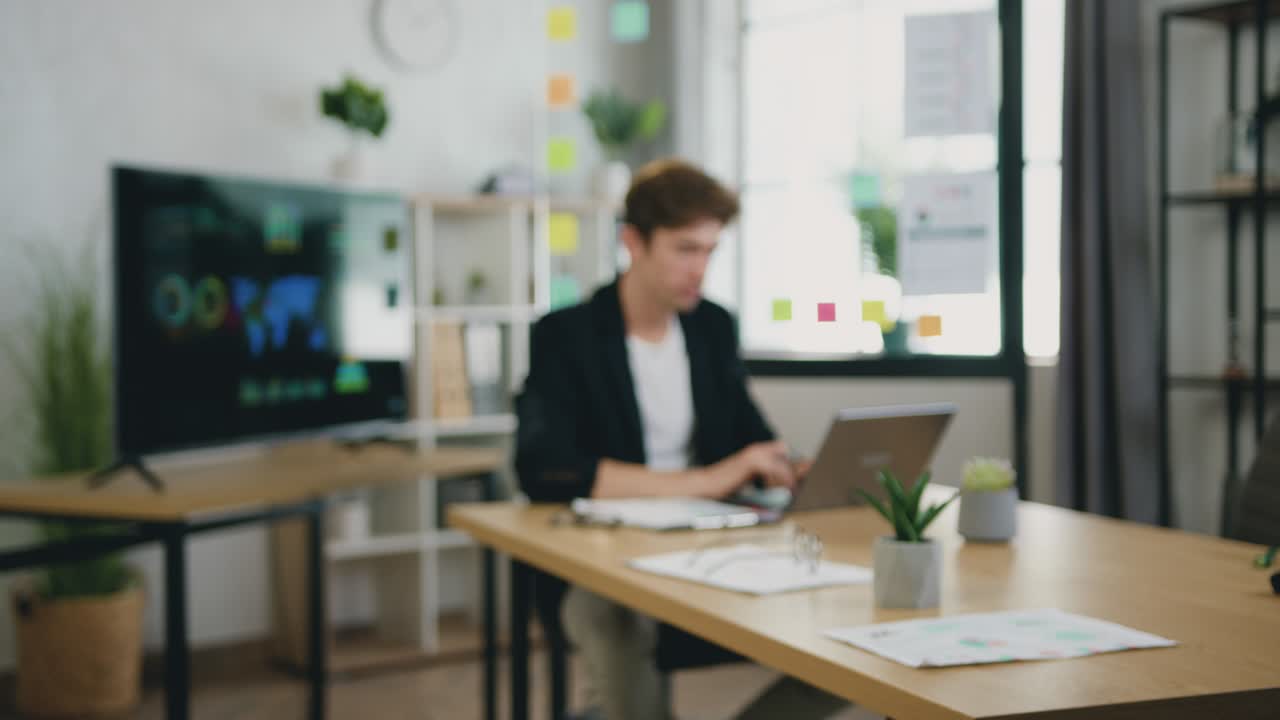 Ritratto di giovane uomo d'affari concentrato che lavora con un computer portatile e documenti cartacei in un ufficio moderno, sorridendo e guardando la fotocamera.