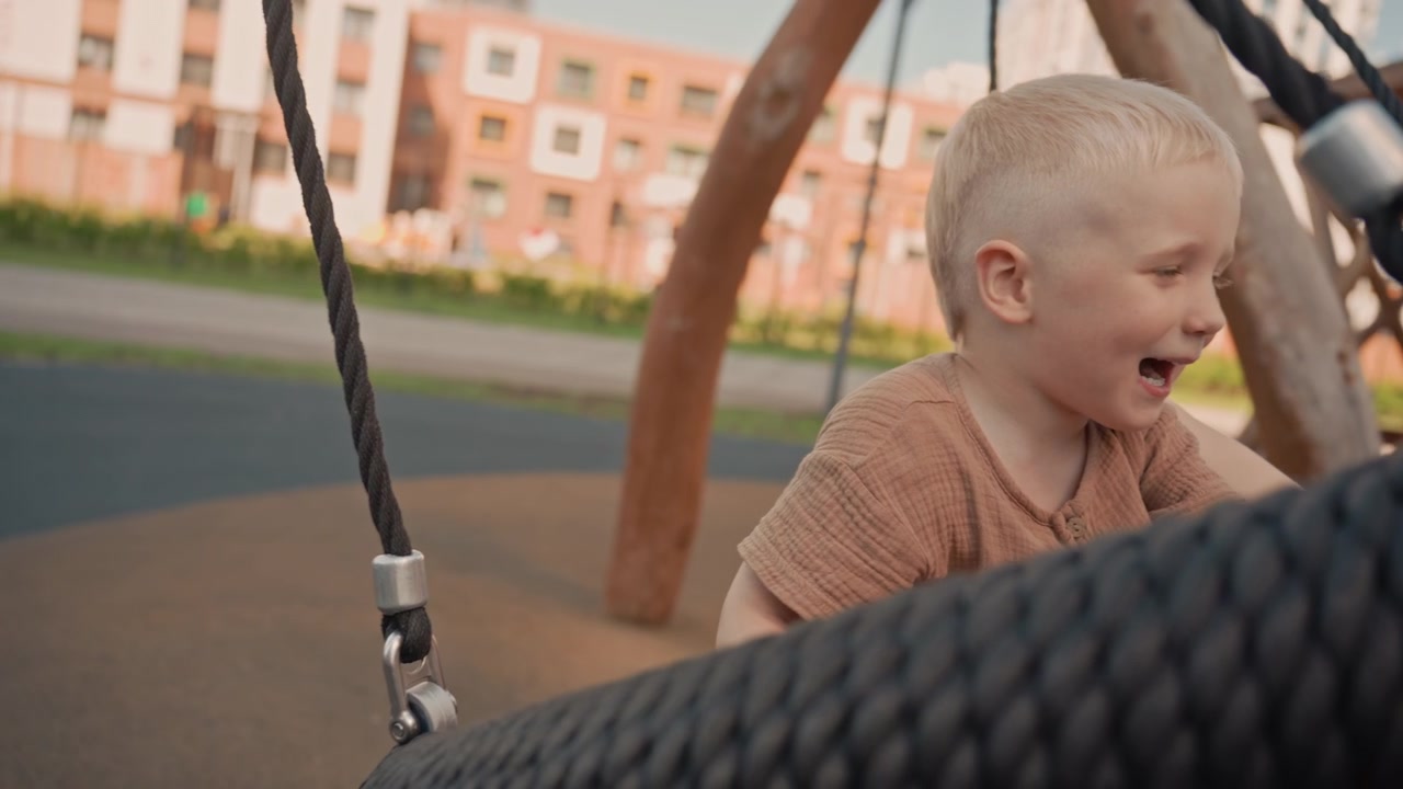 Un bambino si diverte a giocare su un'altalena a pneumatico in un parco giochi all'aperto, muovendosi avanti e indietro con visibile eccitazione. Lo sfondo presenta edifici residenziali in condizioni di luce diurna