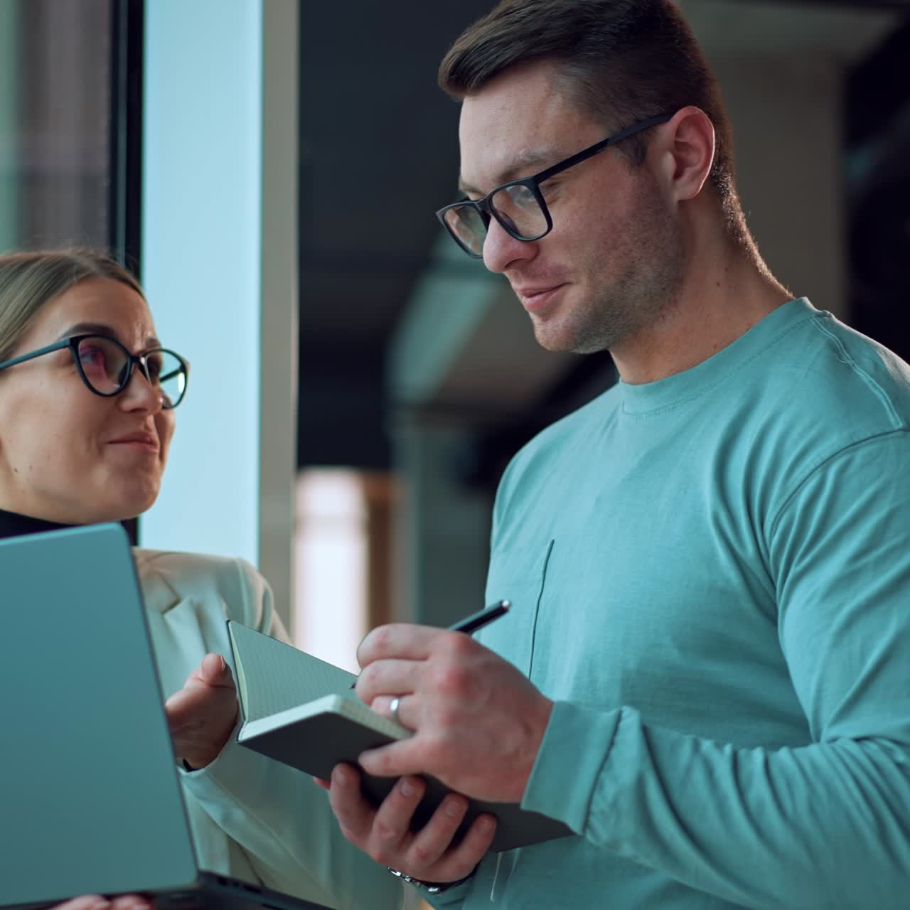 People working together in office communicating standing at the window. Lady holding a laptop and talking to a man taking notes into his paper notebook