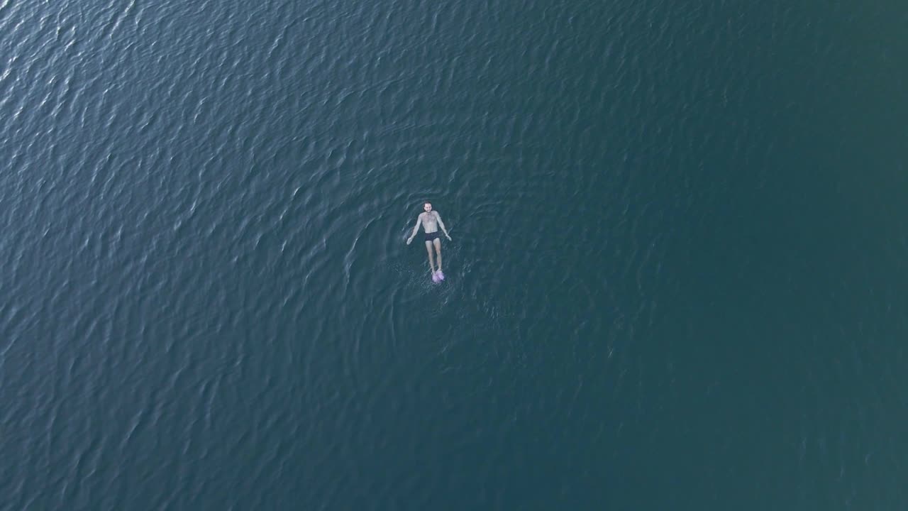 imágenes de drones de arriba hacia abajo de una persona nadando en un lago y haciendo una señal a la cámara