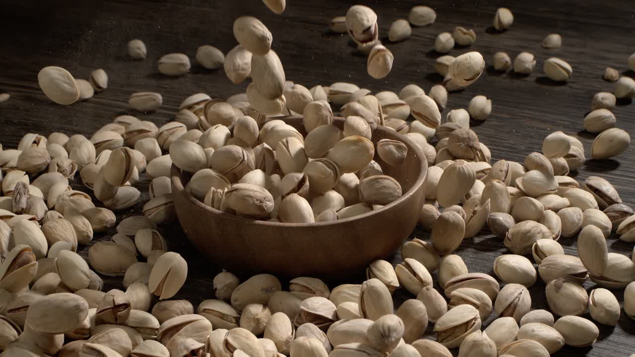 Cuenco de madera lleno de pistachos sobre una mesa de madera oscura; sabrosos frutos secos salados esparcidos alrededor, concepto de snack saludable y alimento natural. Grabado con una cámara superlenta a 1600 FPS.