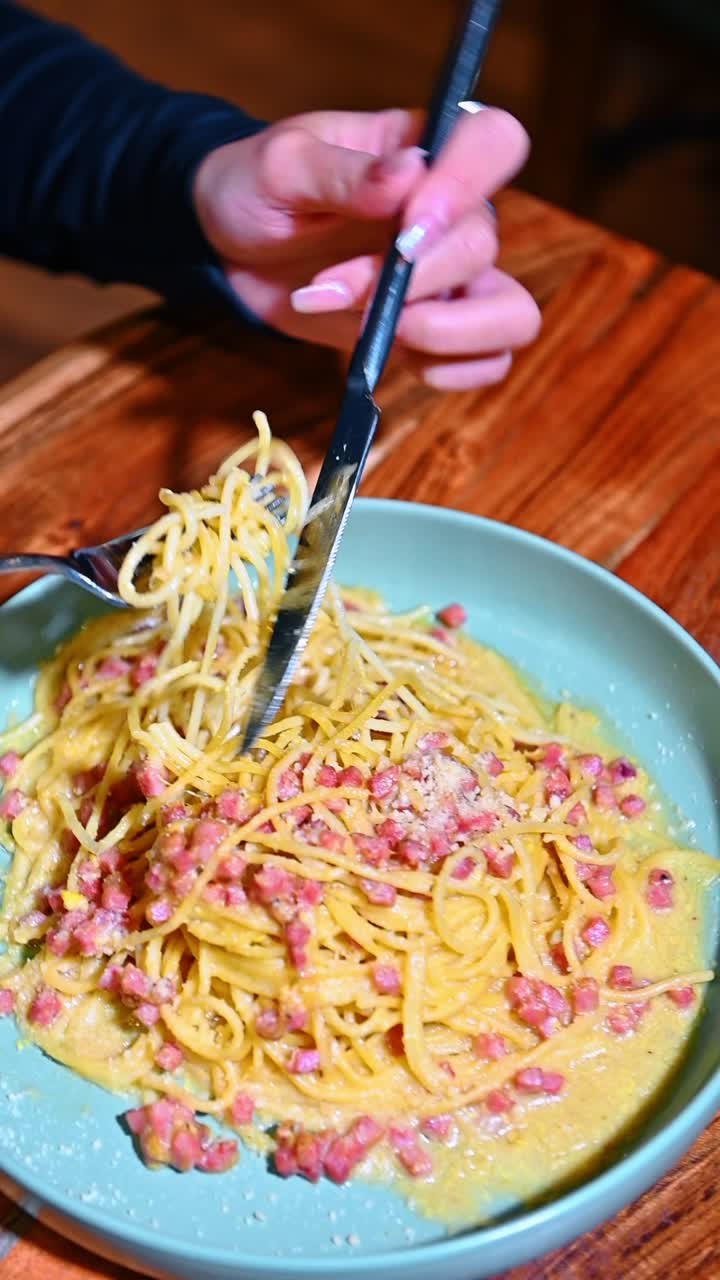 Primo piano delle mani di una donna che mangia pasta italiana al ristorante. Veduta dall'alto di una persona che usa le posate per mangiare spaghetti alla carbonara con pancetta e formaggio.