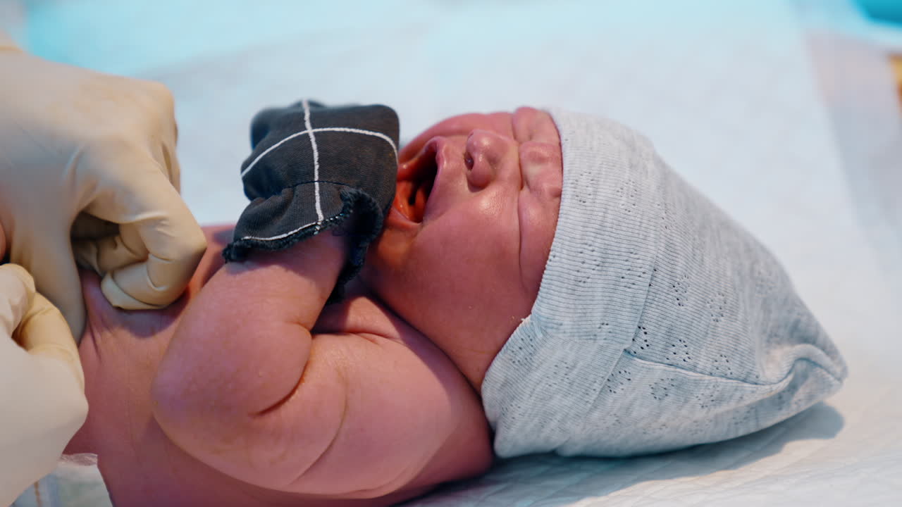 Newborn baby in cap and mittens is screaming bitterly. Medic is putting the diaper on the child. Close up.