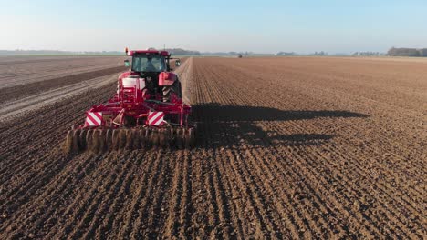 Roter-Traktor,-Der-Braunes,-Erdiges,-Niederländisches-Ackerland-In-Vertikalen-Linien-Unter-Blauem-Himmel-Pflügt