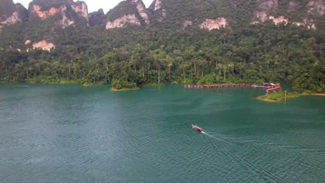 4k drone video panning around a boat sailing around khao sok national park in thailand. the boat is cruising along the beautiful lake with tall cliffs and a dense packed jungle in the background.