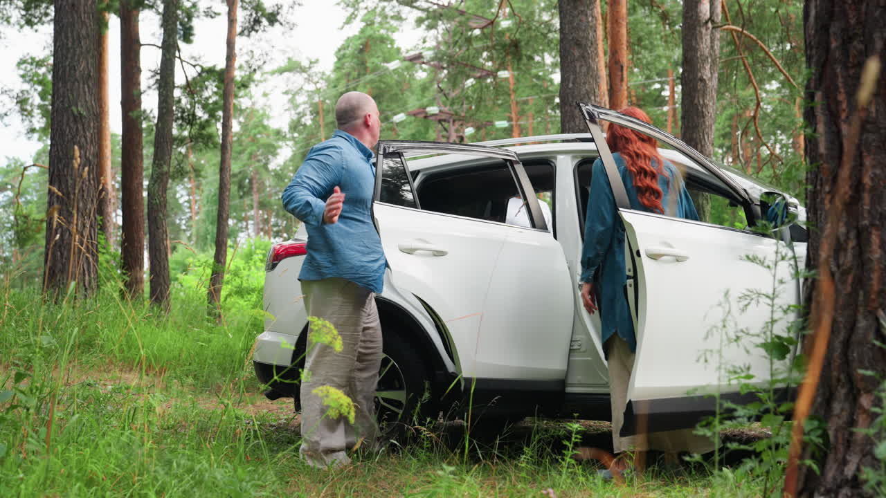 Family suv parked in forest among autumn trees as father helps youngest get into car while other children climb in from opposite door, gentle teamwork during roadside stop, green clearing, open doors
