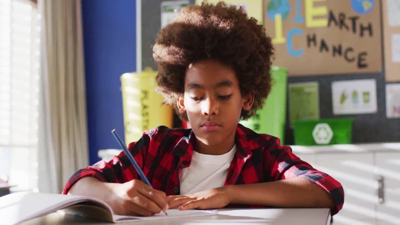 Ritratto di scolaro afroamericano seduto in classe, che prende appunti e guarda la telecamera.
