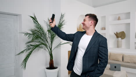 man taking selfie in the big cozy room. male wearing jacket holding the phone above head smiling.