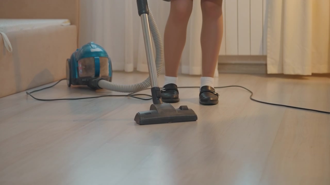 Criança branca aspirando o chão de madeira pela manhã, pequeno aspirador de pó cilíndrico azul, sapatos pretos com meias brancas, barra de saia verde, sofá bege, luz solar suave através das cortinas, movimento de varredura constante, calma.
