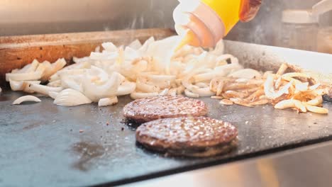 meat for burgers and onions is being cooked on a stove top and oil and spices are being sprinkled on top for seasoning the burger