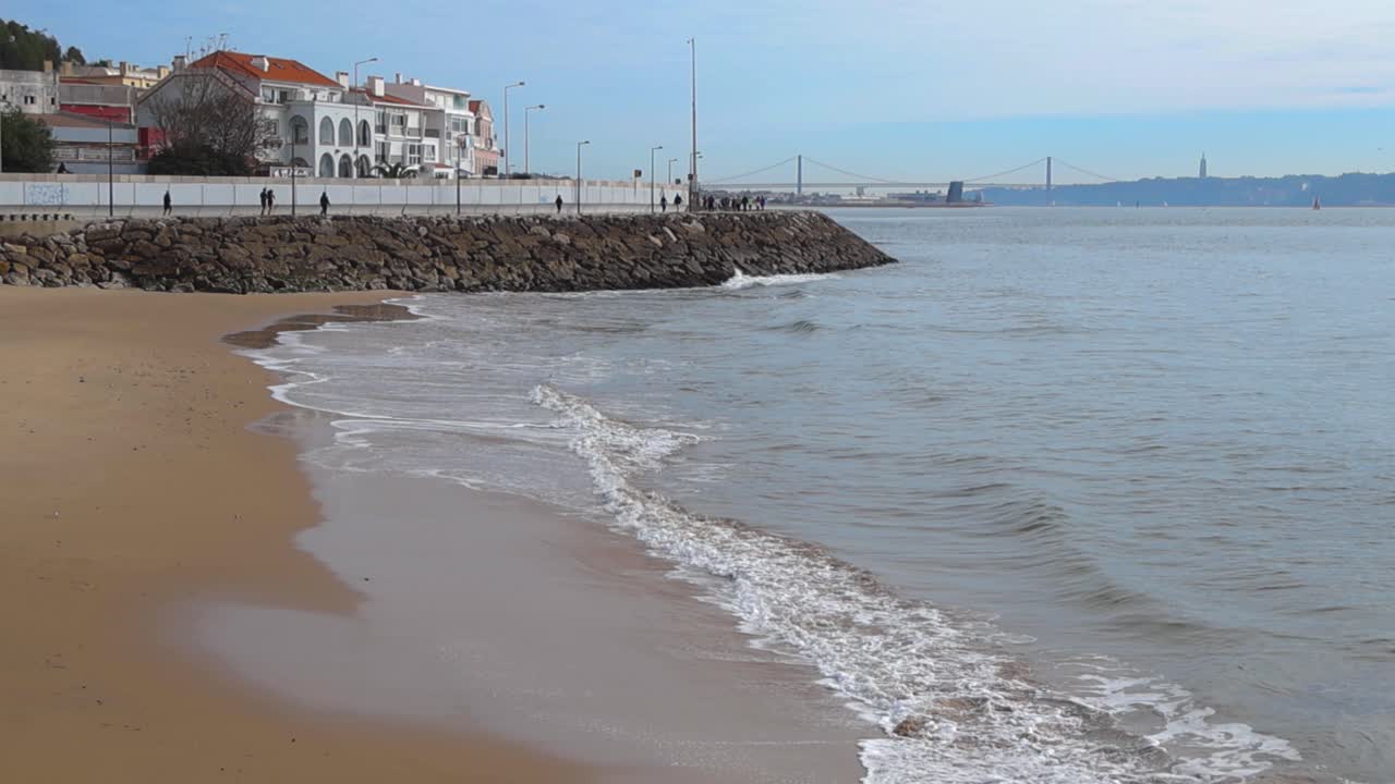 Vista diurna della costa sulla spiaggia di São Bruno con onde leggere che si infrangono sulla sabbia dorata, case bianche e skyline di Lisbona sullo sfondo sotto un cielo sereno