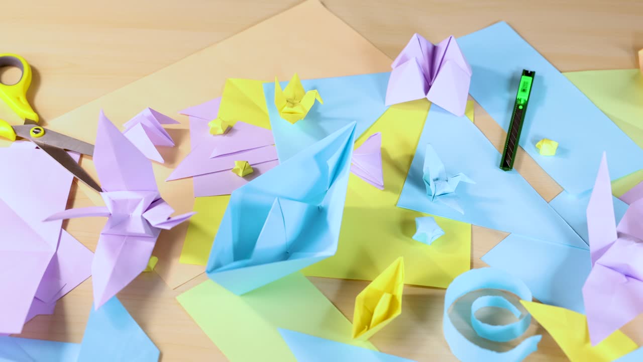 Hands organize pastel origami animals and shapes on wooden desk, overhead view, bright lighting