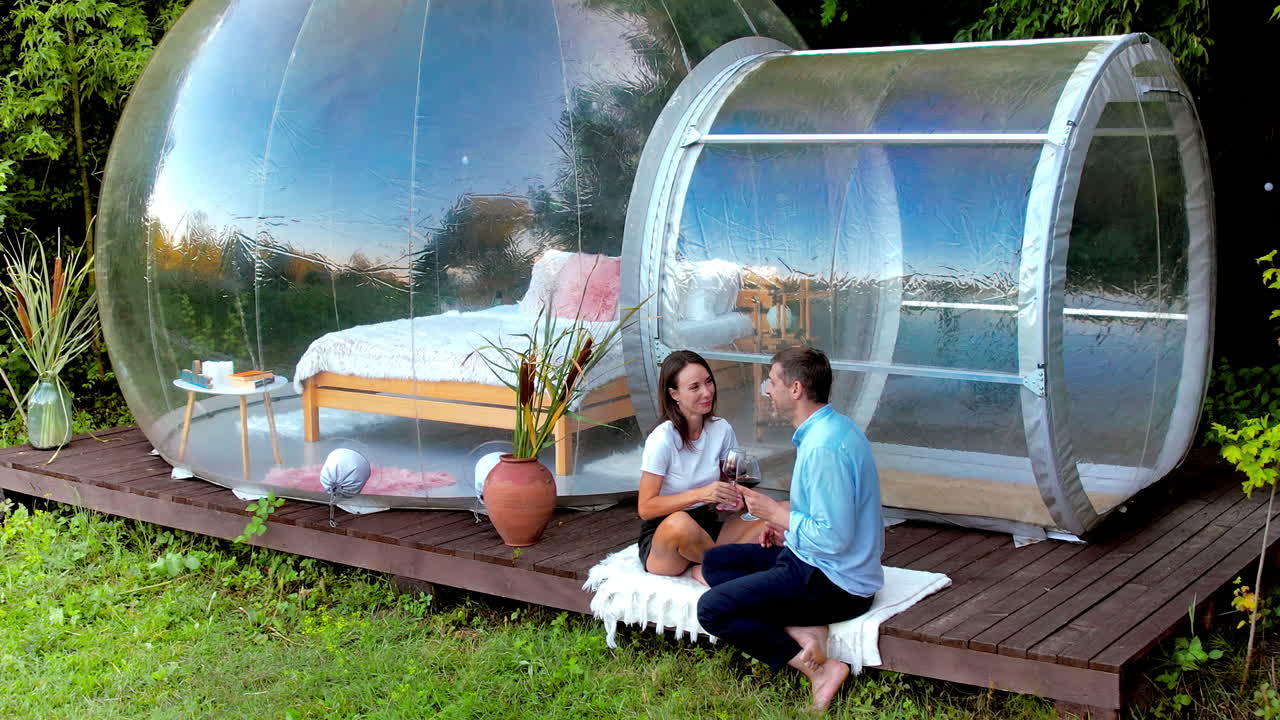 Vista aerea di una coppia felice che beve vino vicino a una tenda a bolla trasparente in un glamping. Natura circostante