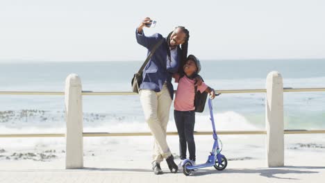 video de un feliz padre e hijo afroamericanos con un scooter tomando una selfie en el paseo marítimo