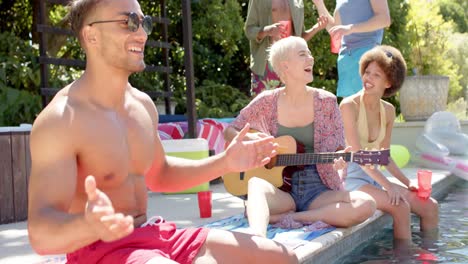 Happy-diverse-group-of-friends-with-drinks-playing-guitar-at-pool-party-in-summer