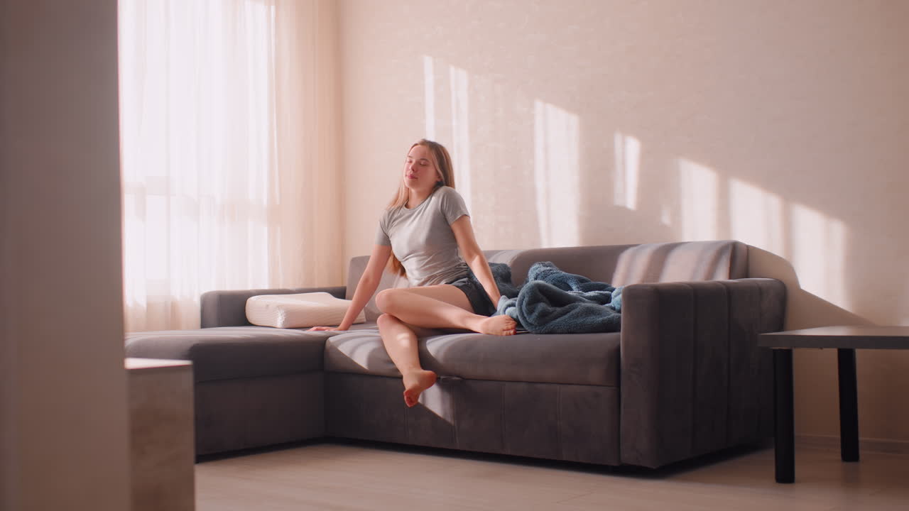 Young girl wakes up on couch stretching muscles after sleep, removing blanket while gazing out window with warm morning sunlight filling cozy room creating peaceful atmosphere of rest