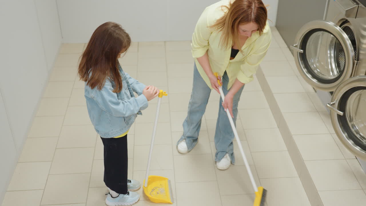High angle view shows girl in denim holding stick dustpan for mother while mother sweeps tiled laundromat floor with broom, teamwork cleaning routine, family help near stainless washers