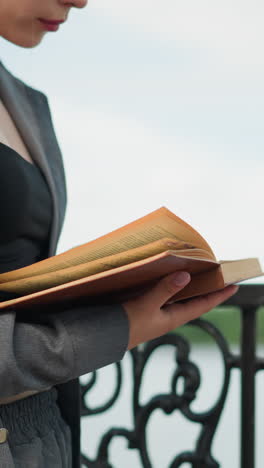 dama sosteniendo un libro abierto en su mano con la mano derecha en las páginas mientras la mano izquierda apoya debajo, el viento sopla suavemente las páginas, con un fondo borroso con cuerpo de agua y vegetación en la distancia