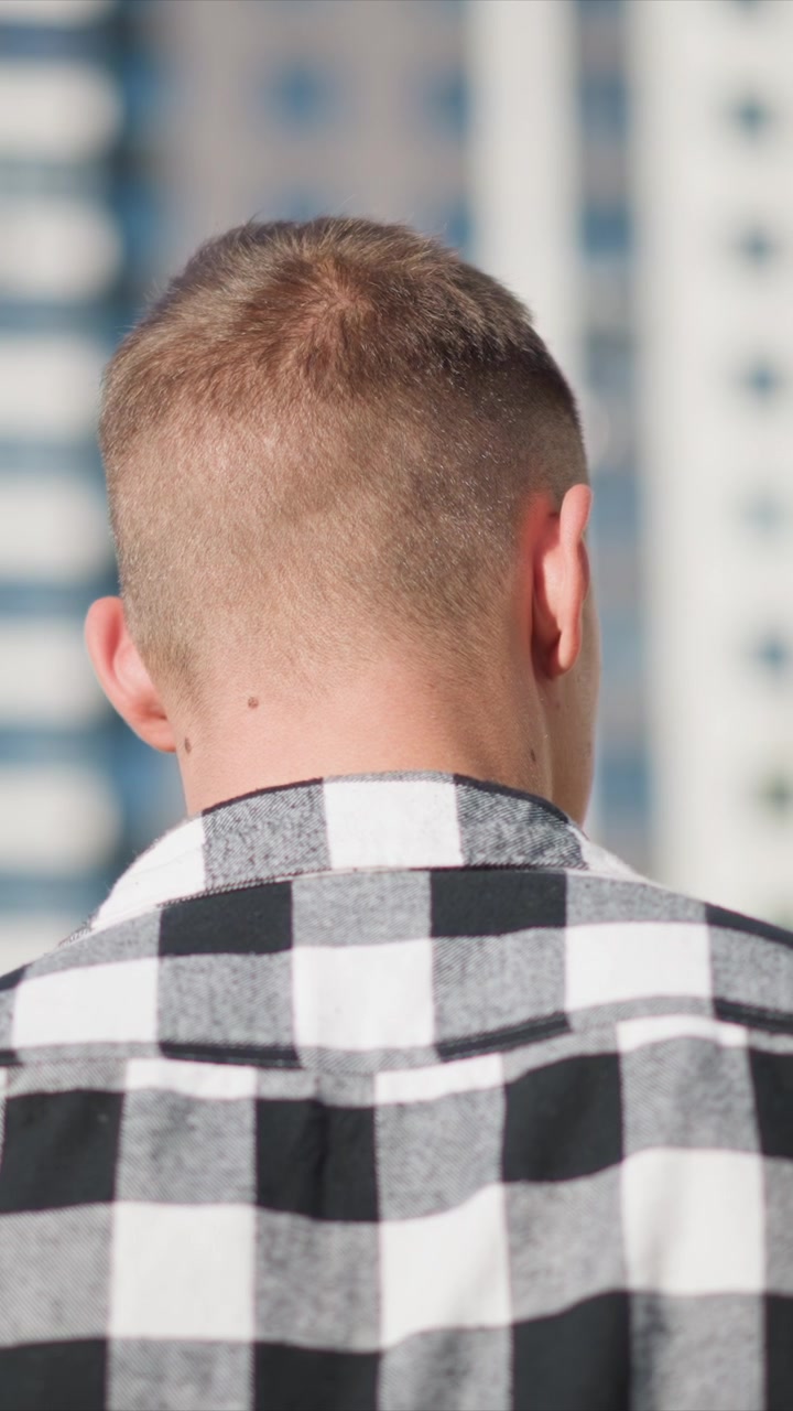 Close up of man with short hair wearing black and white checkered shirt seen from behind. background shows urban setting with tall buildings visible through soft daylight lighting. scene