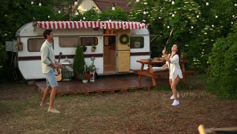 Two-young-tourist-couple-play-badminton-next-to-their-trailer-in-american-retro-style.-Active-vacation,-rest,-traveling.-Summer,-warm-day