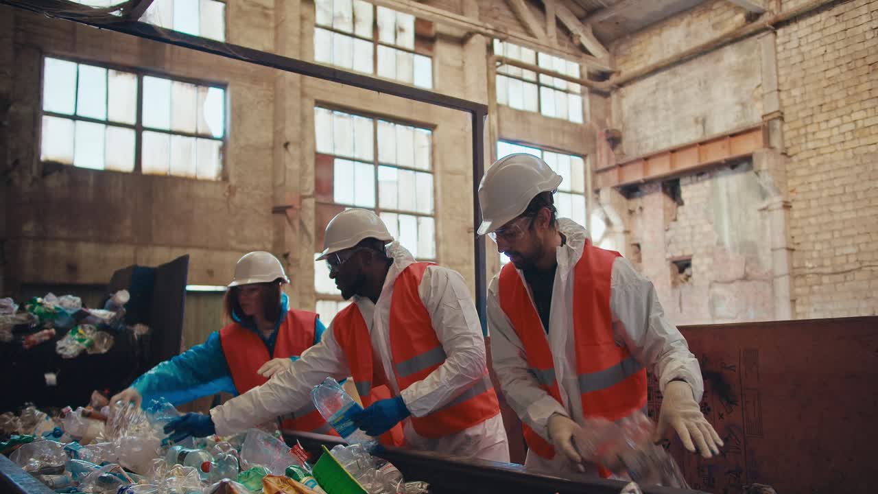 A trio of waste recycling plant workers in special protective uniforms and white helmets stand near the conveyor and lay out bottles depending on the color of the plastic at the plant
