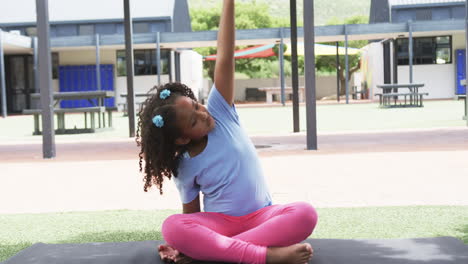 Biracial-girl-with-curly-hair-sits-cross-legged-outdoors-at-a-playground-in-school