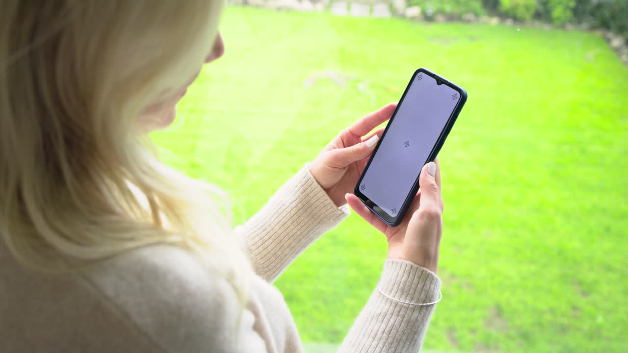 Woman standing by window inspecting phone spinner, angling device toward light and tapping screen