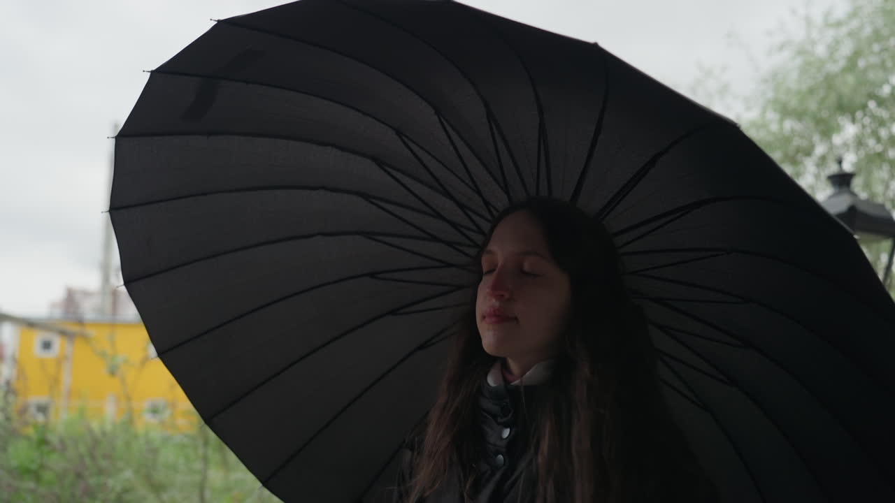 Giovane donna che cammina sotto un ombrello nero, parco cittadino sullo sfondo. Giornata nuvolosa con pioggerellina, lampione ed edificio giallo visibili, capelli lunghi e cappotto, espressione riflessiva, inquadratura a ritmo lento che cattura il momento
