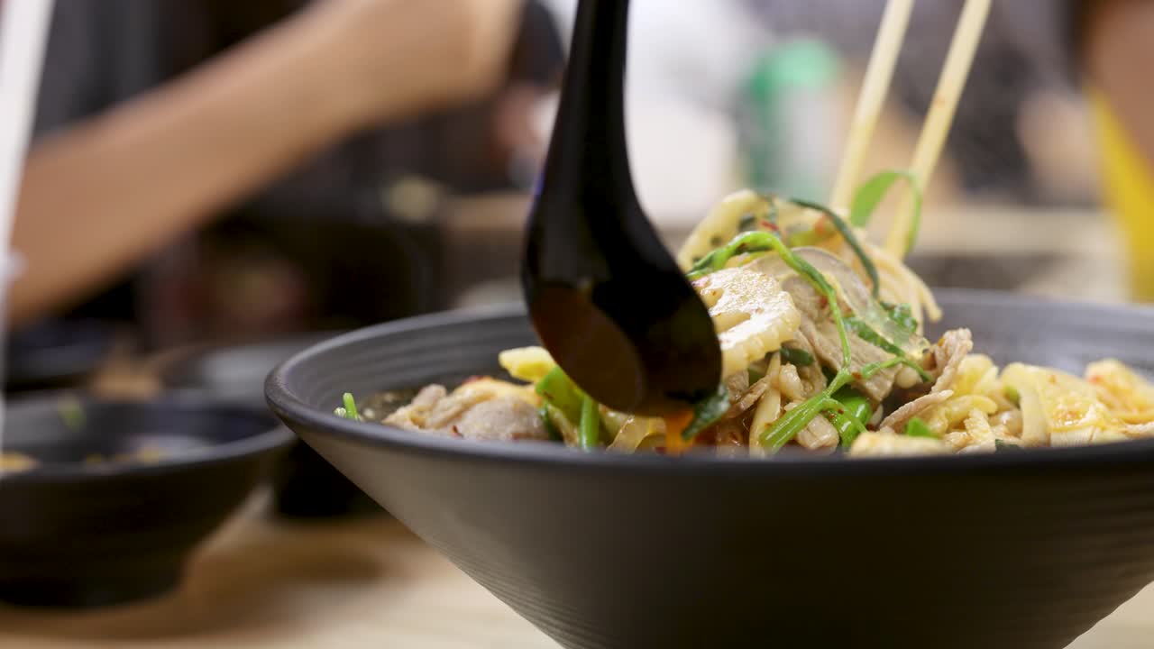 Hand stirs hearty Asian hotpot noodles, tofu, vegetables in savory broth under bright lighting
