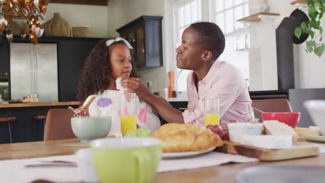 Video-of-happy-african-american-mother-wiping-daughter's-face-at-family-breakfast-table