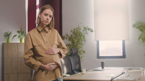 Mujer-Embarazada-Posando-En-Cámara-Mientras-Toca-Su-Vientre-En-La-Oficina