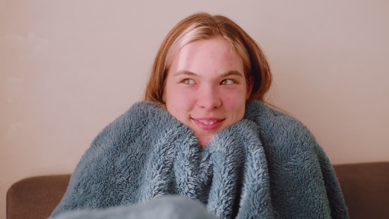Elegant lady seated on sofa feeling cold covering herself with fluffy blanket looking in funny way as sunlight passes through curtain creating cozy indoor atmosphere of warmth and comfort
