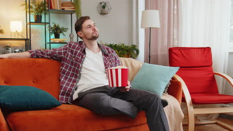 happy man eating popcorn watching interesting tv serial sport game online movie sits on home sofa