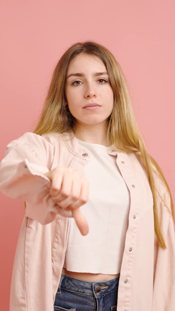 Young woman expressing disapproval with thumbs down on pink background