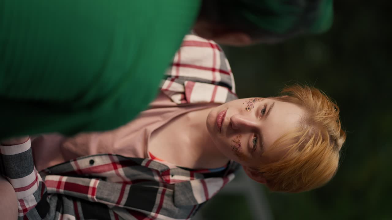 Over the shoulder: vertical video a blonde girl with a short haircut and sparkles on her face in a plaid shirt looks intently at her girlfriend with a green haircut in a Green shirt and adjusts her makeup on her date in the park