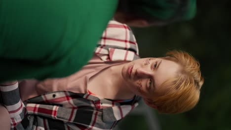 over the shoulder: vertical video a blonde girl with a short haircut and sparkles on her face in a plaid shirt looks intently at her girlfriend with a green haircut in a green shirt and adjusts her makeup on her date in the park