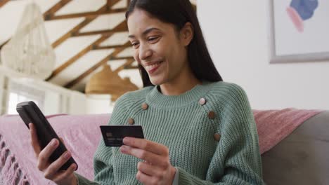 biracial woman using smartphone and holding credit card in living room