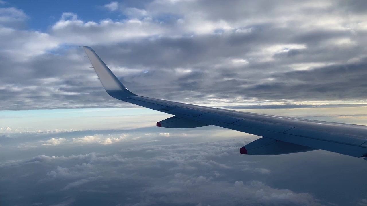 Ala di aereo sopra nuvole sparse durante il volo, con strati di nuvole grigie sopra e squarci di cielo blu